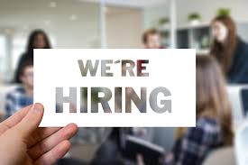 Now Hiring sign on a blurred background of an office space.